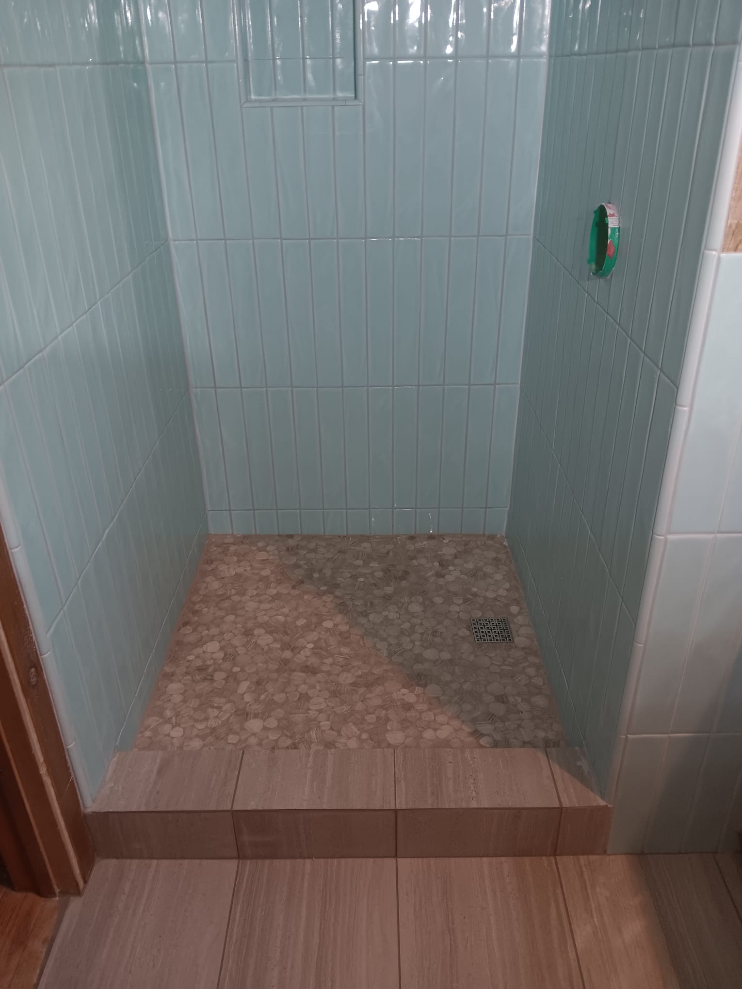 Shower in pale aqua tile stacked vertically with a pebble floor and a wood-look porcelain curb