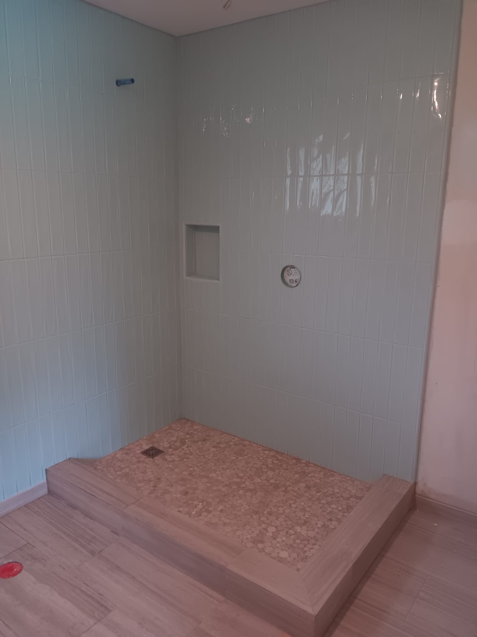 Shower in white gloss tile stacked vertically, with a recessed niche, pebble pan and wood-look plank curb